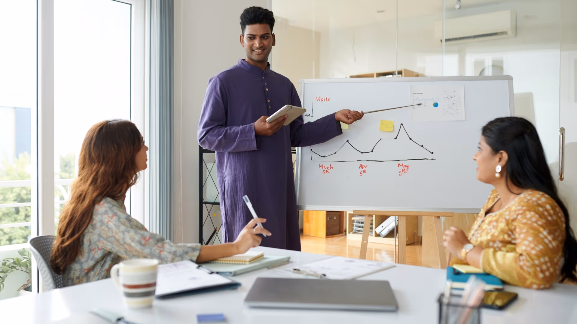 Man in purple kurta presenting data with a pointer to two women seated at a table in a bright office.