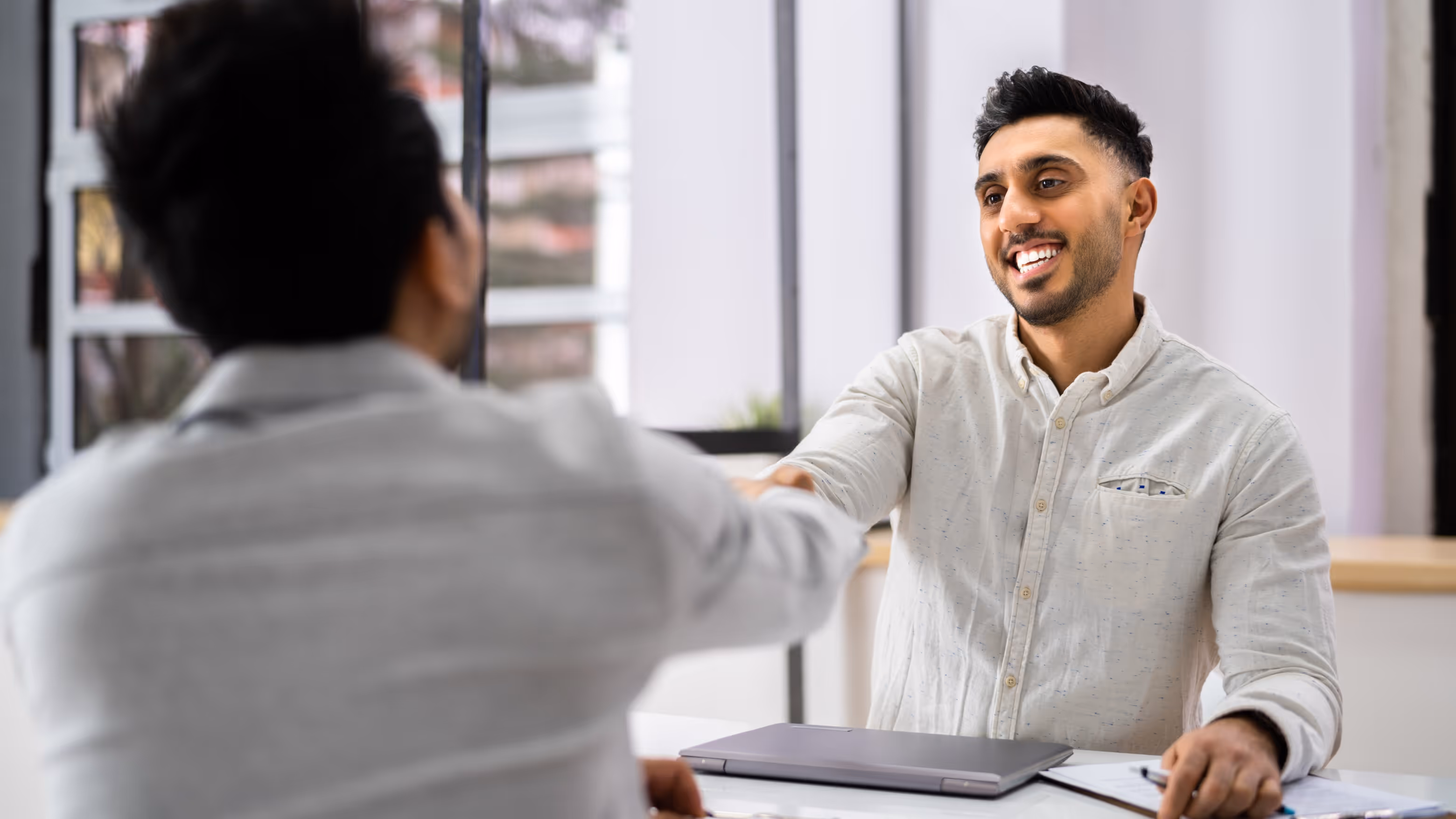 Two men shaking hands across a desk in a bright office, one smiling with a laptop and clipboard on the table.