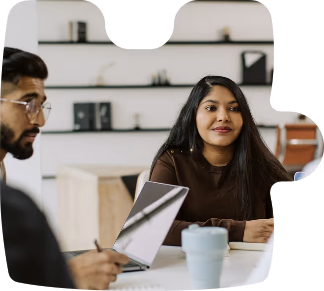 Two people having a discussion at a desk with a laptop and a cup in a modern office setting.