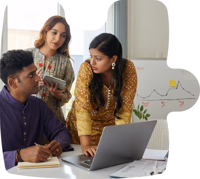 Three colleagues discussing work around a laptop, with one taking notes and a whiteboard with a graph in the background.