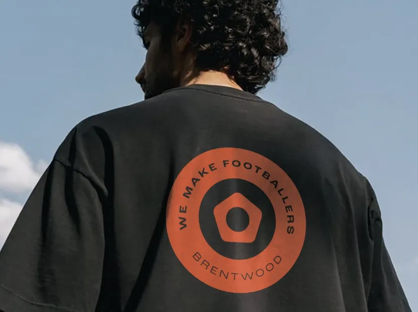 Man with curly hair wearing a black t-shirt with an orange circular logo reading 'WE MAKE FOOTBALLERS BRENTWOOD' on the back against a blue sky.