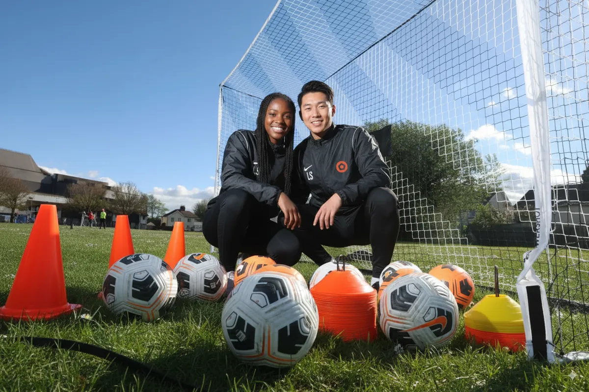 Two soccer coaches kneeling on grass in front of a goal net with training cones and balls around them under a clear blue sky.