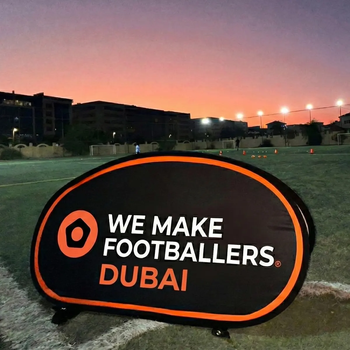 Black and orange pop-up banner on a soccer field at dusk with the text 'WE MAKE FOOTBALLERS DUBAI'.