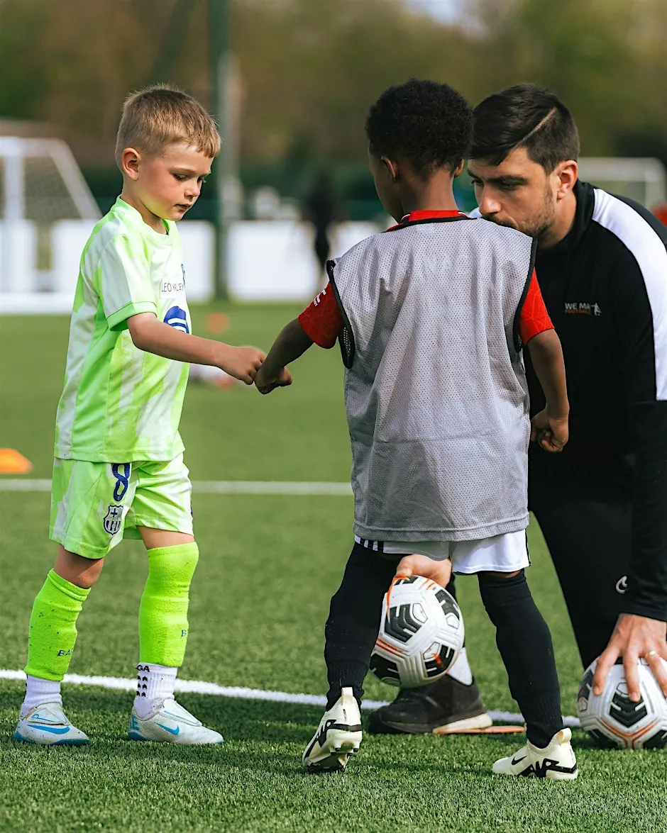 Youth soccer coach crouching and holding a soccer ball while two young boys shake hands on a soccer field.