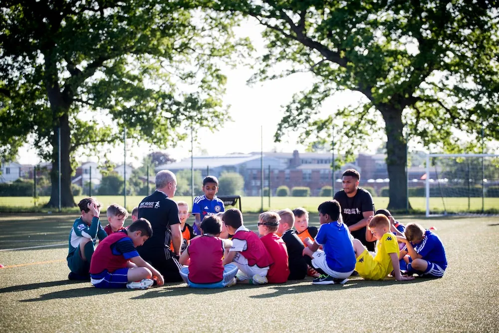 Children sitting in a circle on a soccer field listening to two coaches under large trees.