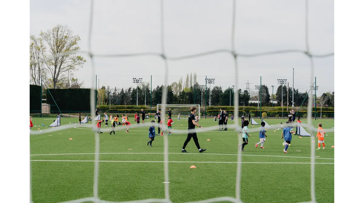 Children playing soccer on a green field with coaches guiding them, seen through a goal net.