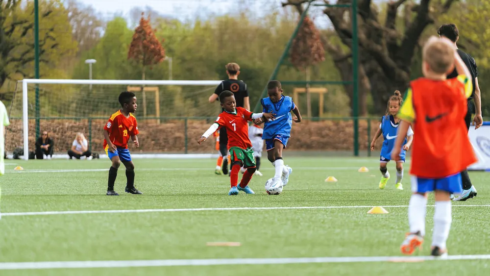 Children playing soccer on a green field with a goalpost and trees in the background.