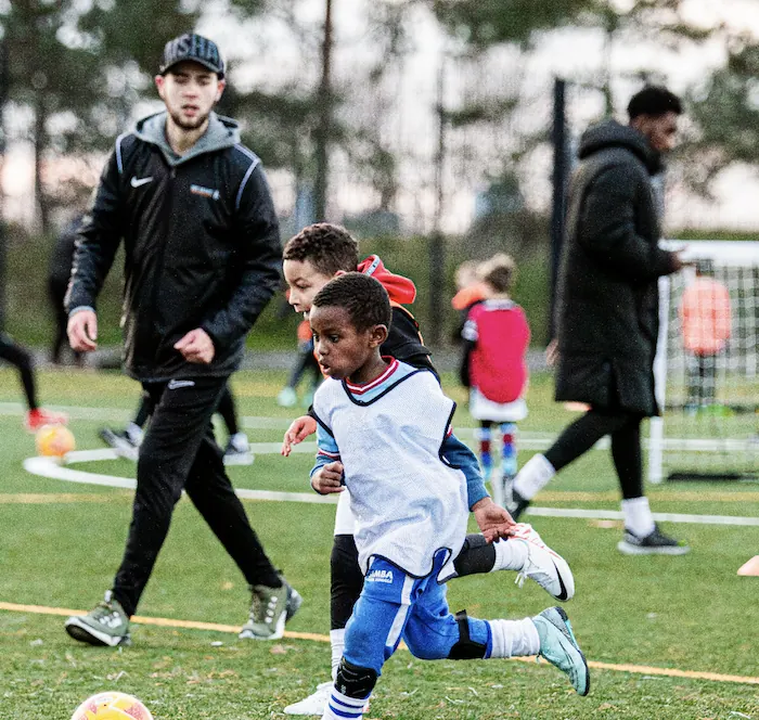 Two young boys playing soccer actively on a green field with two coaches observing in the background.