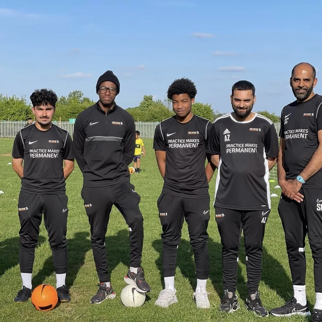 Five men standing on a grassy field with two soccer balls, wearing black sportswear with 'Practice Makes Permanent' text.