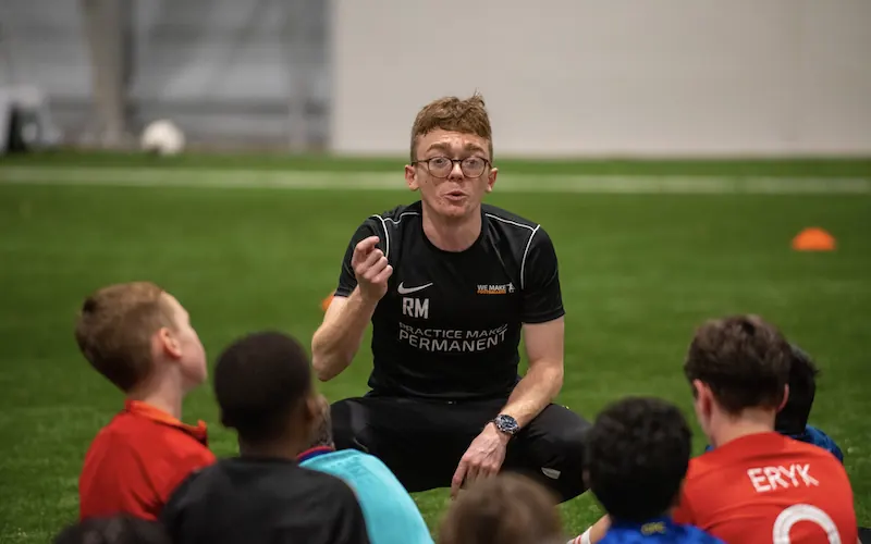 Coach with glasses and black sportswear talking to a group of children sitting on indoor soccer field.