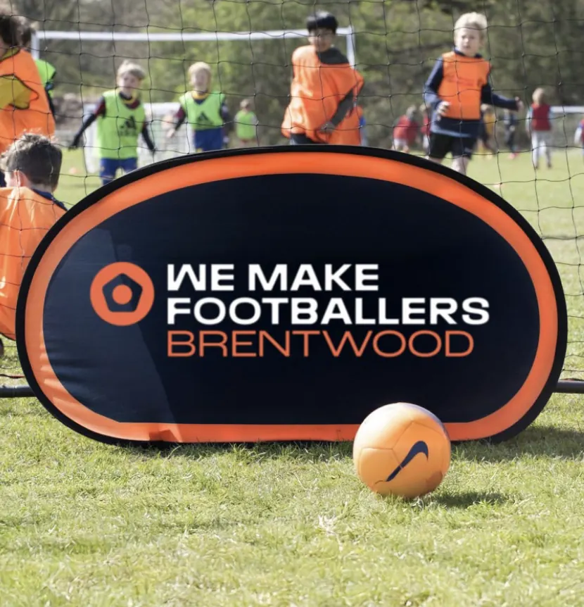 Young children playing football on a field behind a small black and orange goal with the text 'We Make Footballers Brentwood' and an orange football with a black Nike swoosh in front.