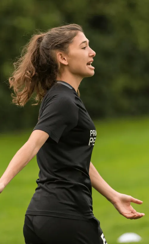 Female soccer player in black uniform with ponytail, gesturing with arms while talking on a green field.