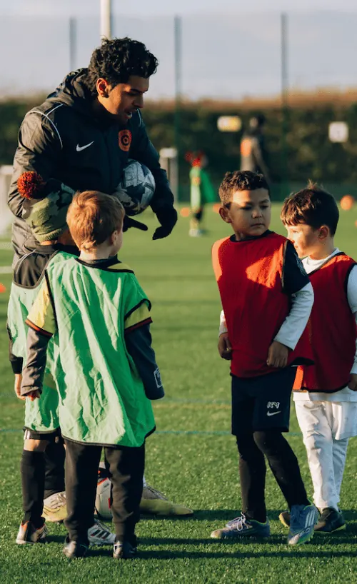Youth soccer coach giving instructions to young players wearing red and green training vests on a grassy field.