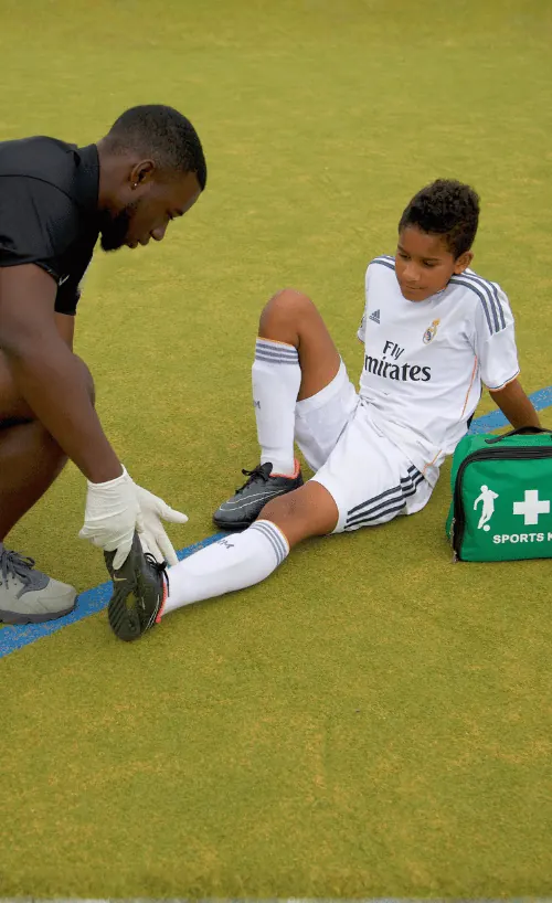 A male sports medic wearing gloves assists a young soccer player in a white uniform sitting on the grass, holding the player's foot.