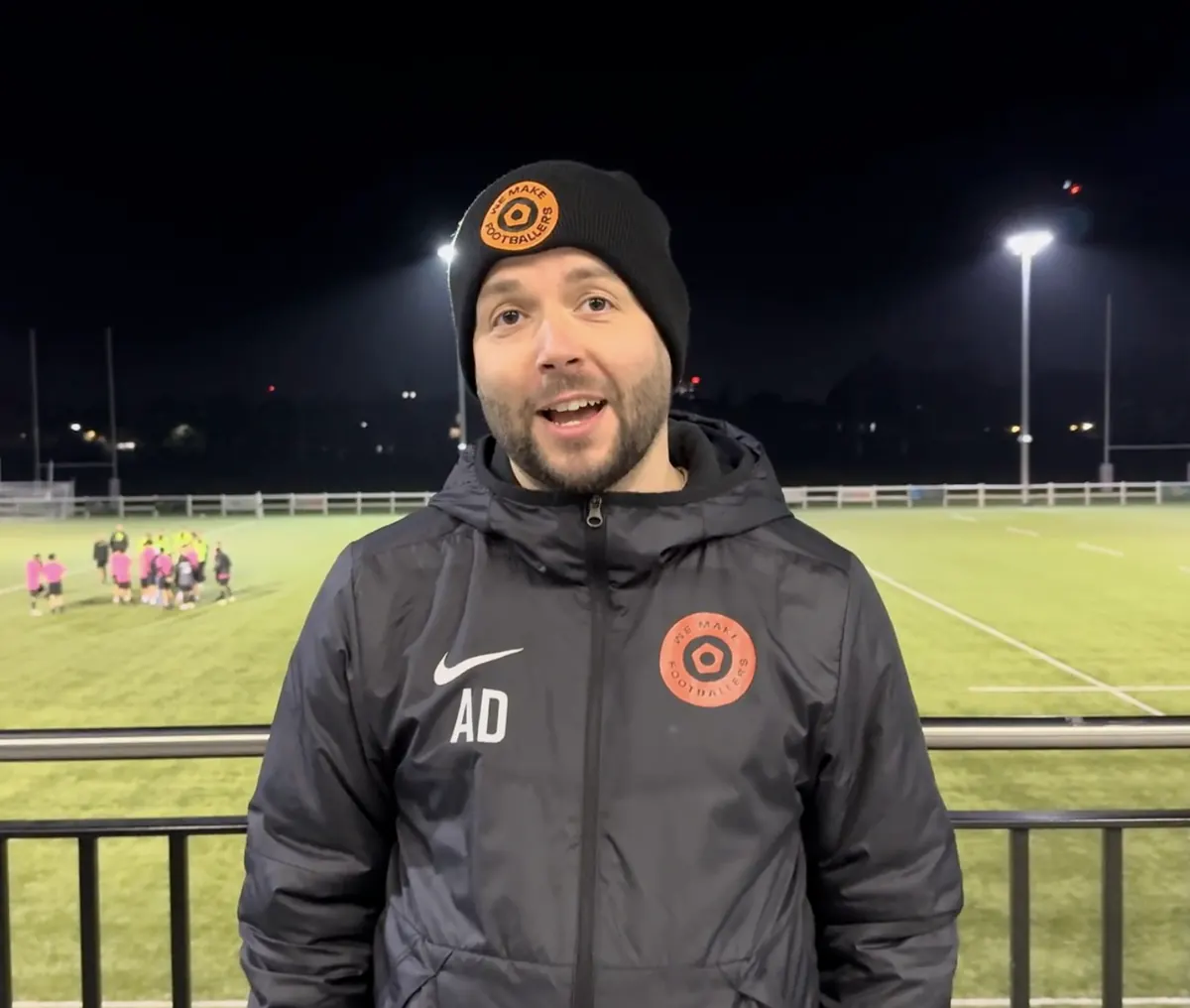 Man wearing a black beanie and jacket with 'We Make Footballers' logo standing on a sports field at night.