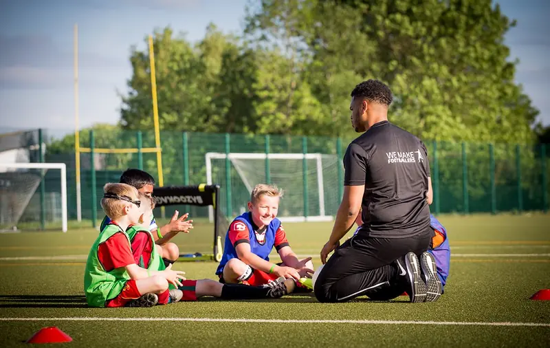Youth soccer coach kneeling and instructing four children sitting on a grassy field with soccer goals in the background.