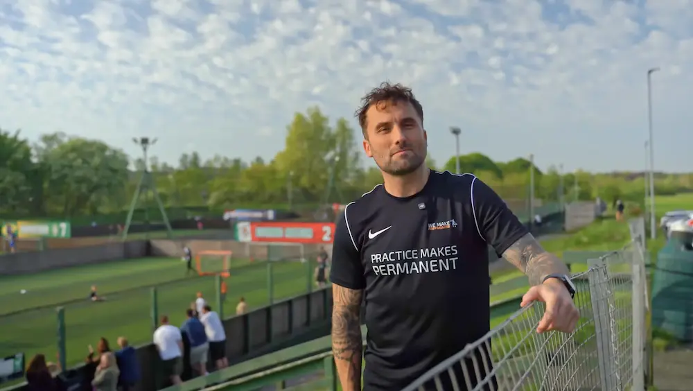 Man with tattoos wearing a black shirt that says 'Practice Makes Permanent' leaning on a fence at an outdoor sports field with people in the background.