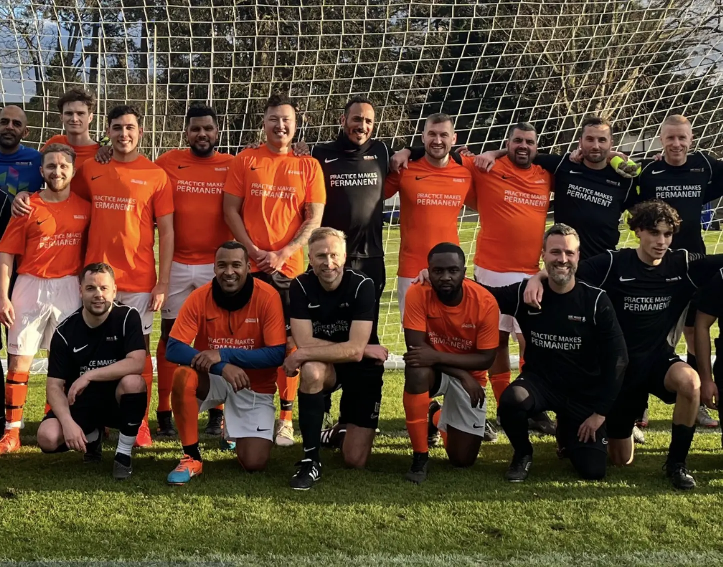 Two soccer teams posing together on a field in front of a goal net, wearing orange and black jerseys that say 'PRACTICE MAKES PERMANENT'.