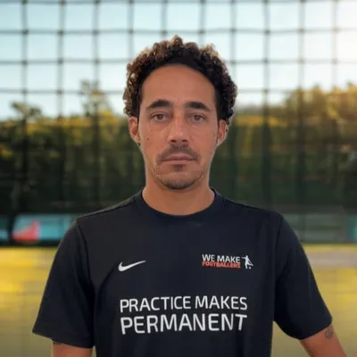 Man wearing a black Adidas jacket with 'DP' and 'We Make Footballers' logos standing inside an indoor soccer facility.
