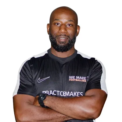 Confident man with a beard wearing a black sports shirt with text 'We Make Footballers' and 'Practice Makes' posing with arms crossed.