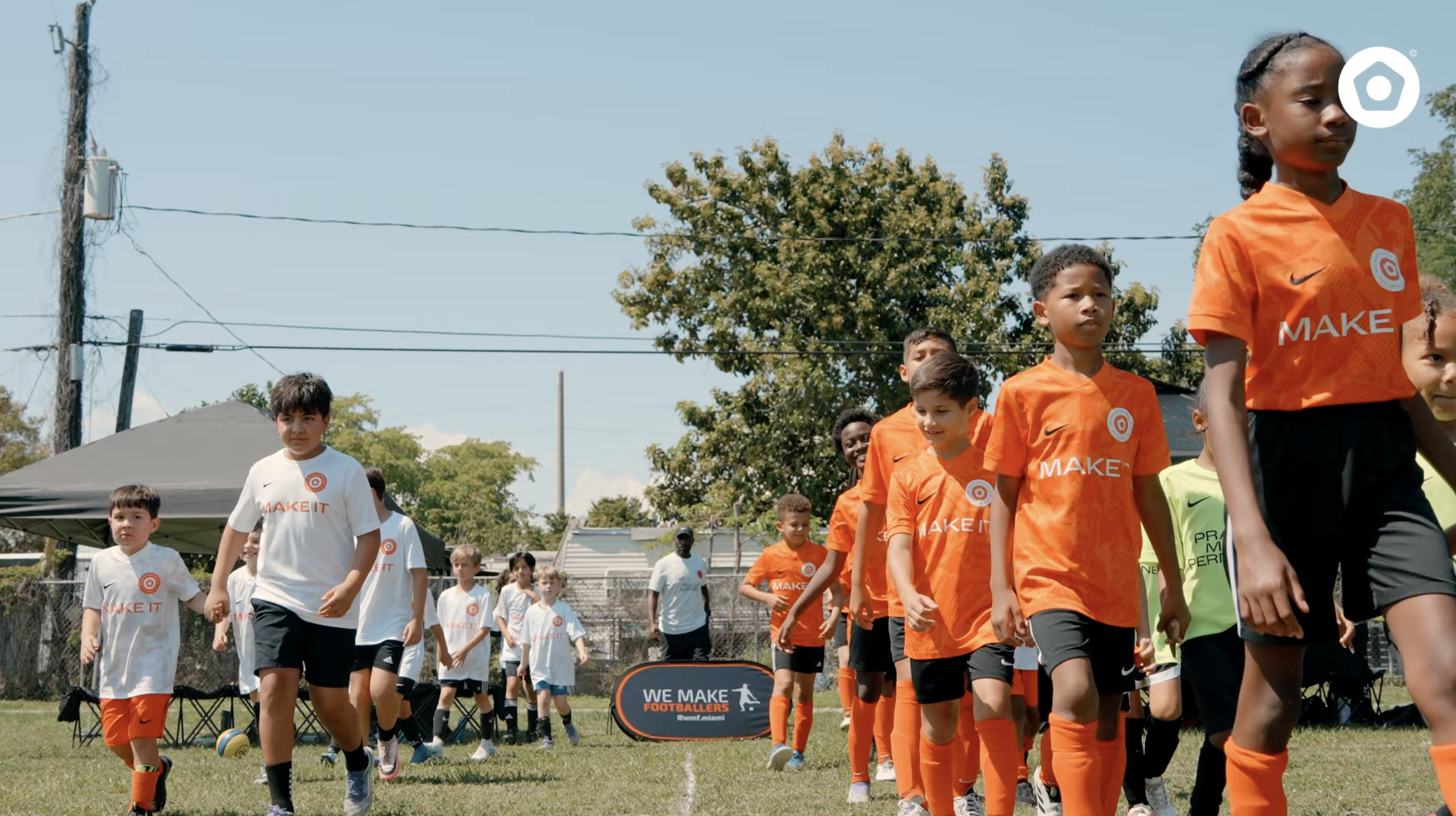 Children walking on a soccer field wearing orange and white 'MAKE IT' jerseys on a sunny day.