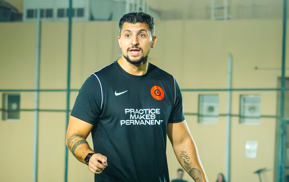 Young girl in neon soccer uniform dribbling a soccer ball around an orange cone with coach crouching nearby giving instructions on a grassy field.