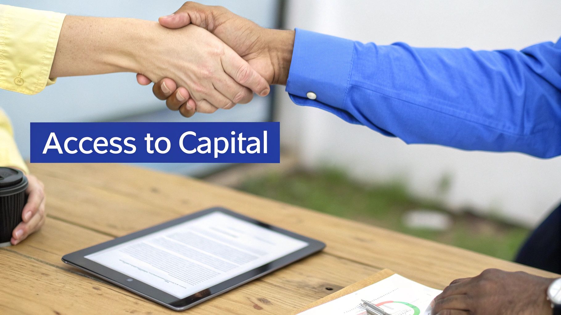 Two business people shake hands over a wooden table with a tablet, symbolizing 'Access to Capital'.