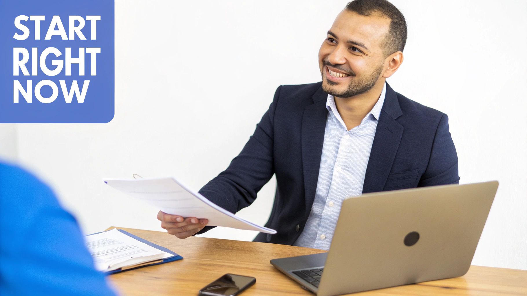 A smiling man in a suit hands documents to another person during a business meeting, with a laptop on the desk.