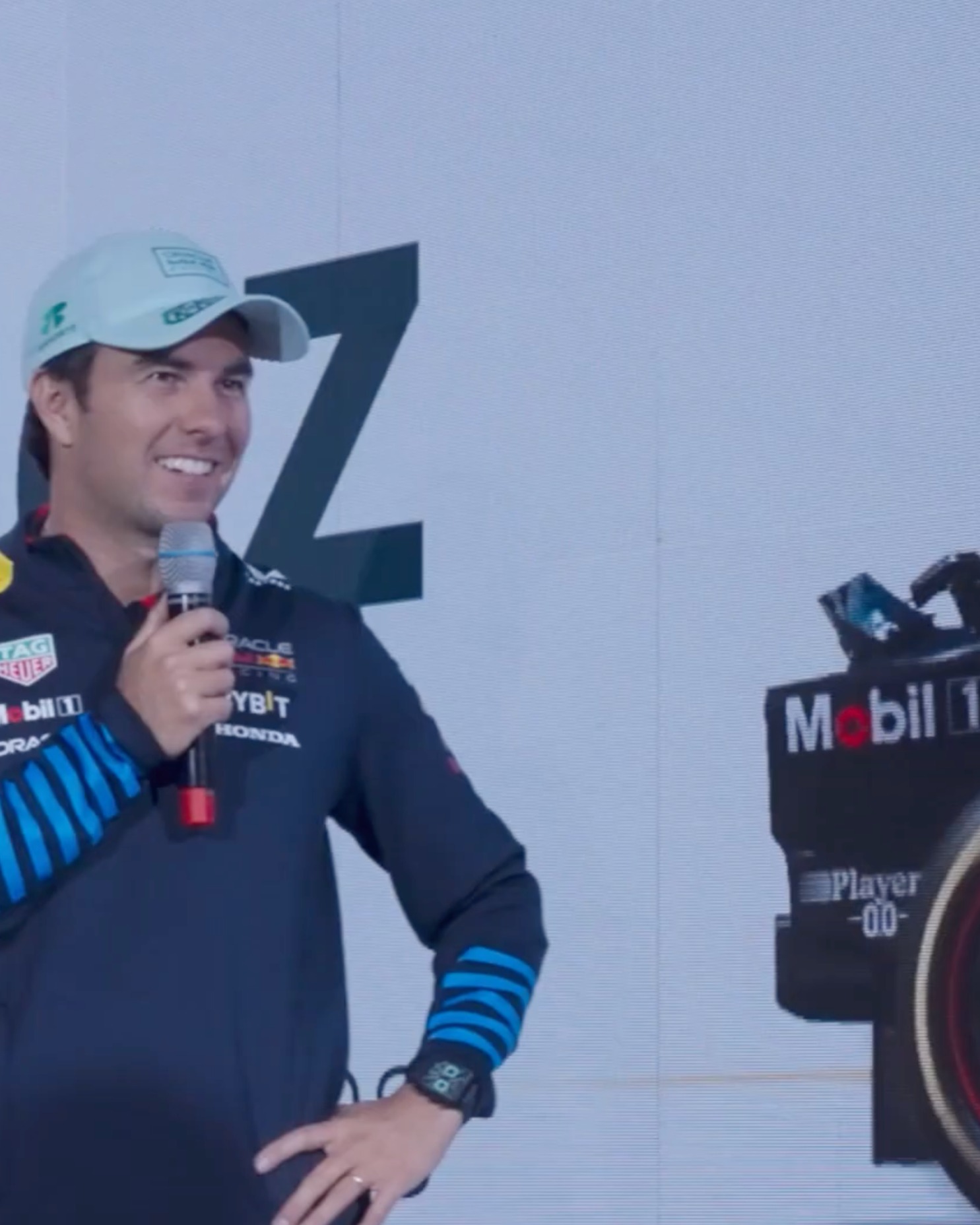 Smiling man in a Dark Blue Red Bull Racing jacket and white cap holding a microphone during a presentation with a Formula 1 car to his right.