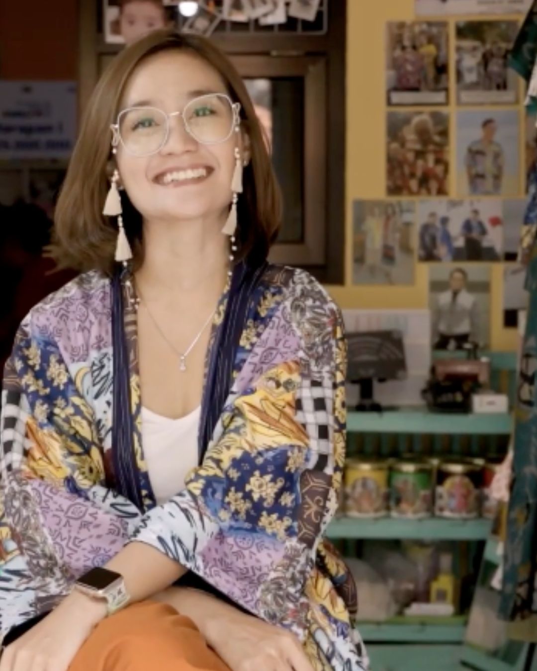Smiling woman with glasses, wearing a colorful patterned jacket and earrings, sitting in a room with clothing and photos on the wall.