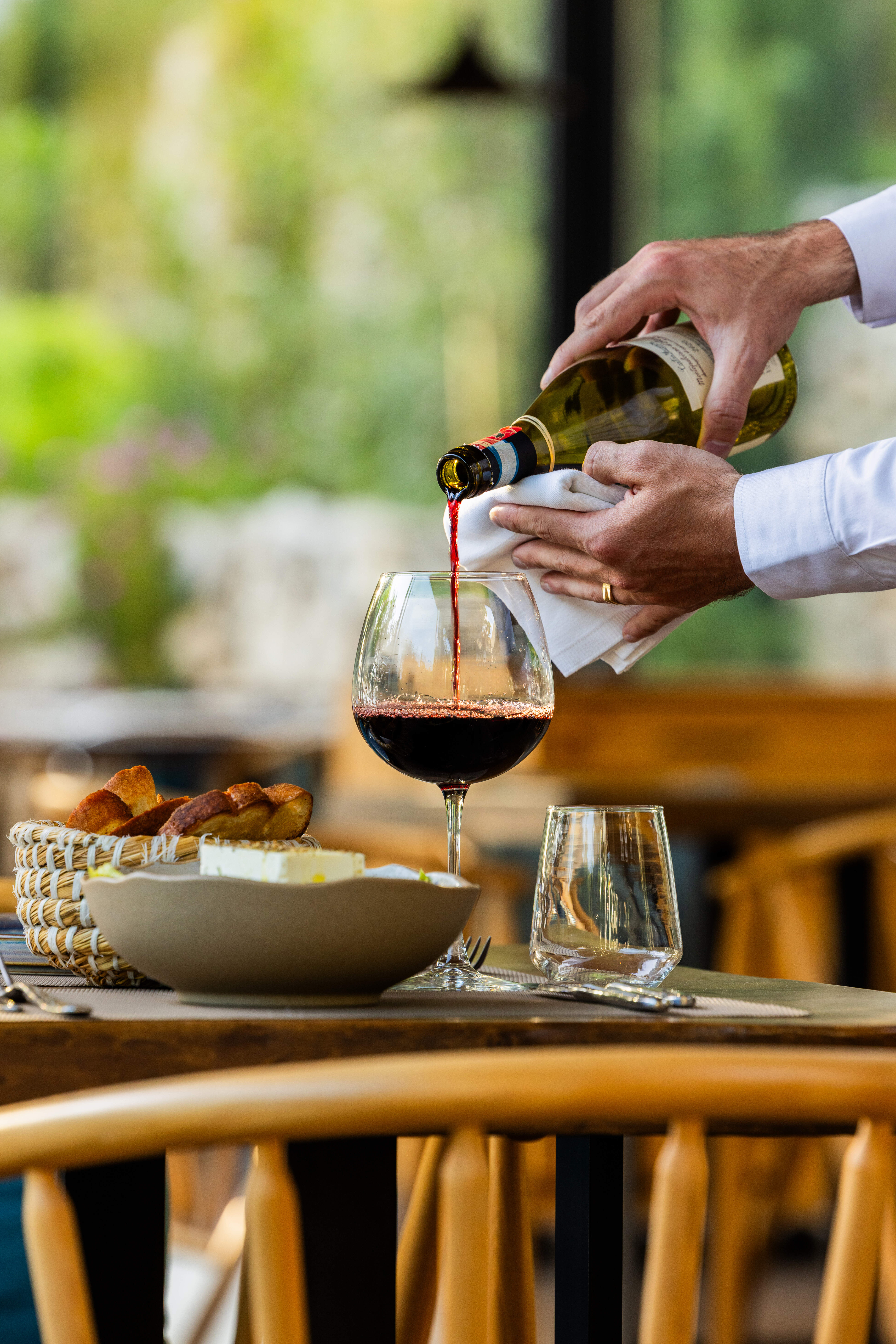 Hands pouring red wine from a bottle into a glass on a table with a basket of bread and a bowl of cheese.