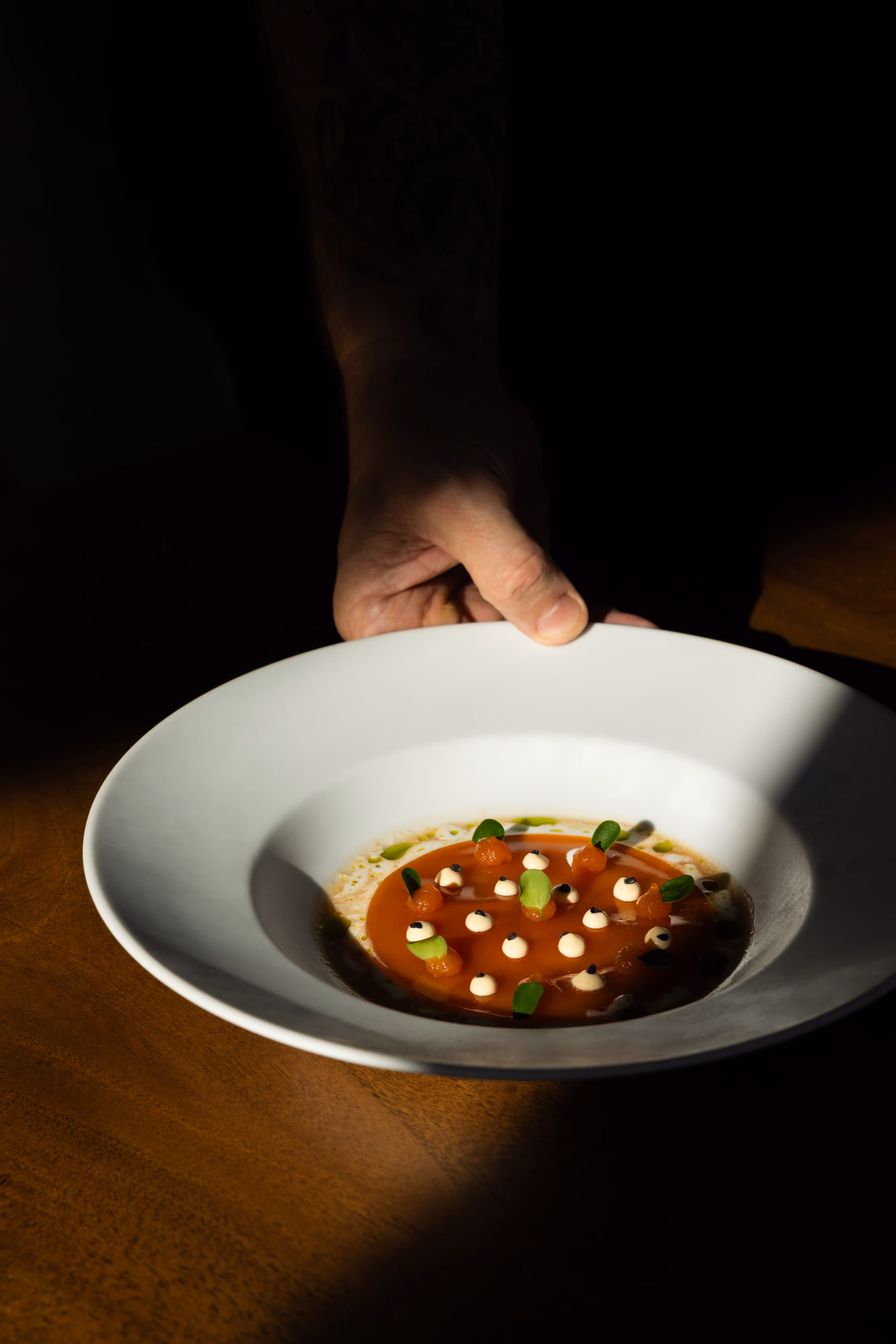 Hand holding a white bowl with a gourmet tomato soup garnished with small cream dollops, green leaves, and small spheres.