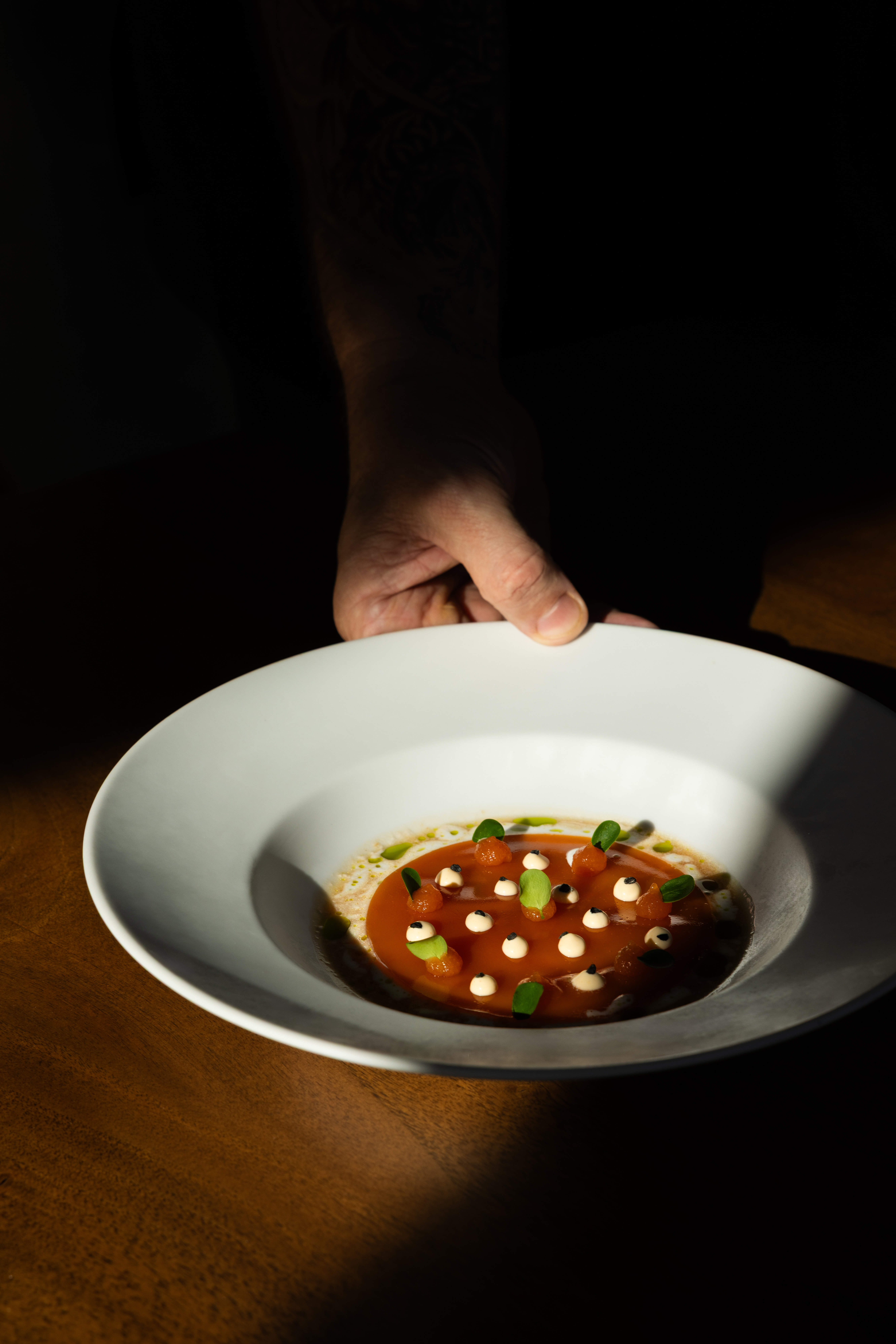 Hand holding a white bowl with a gourmet tomato soup garnished with small cream dollops, green leaves, and small spheres.