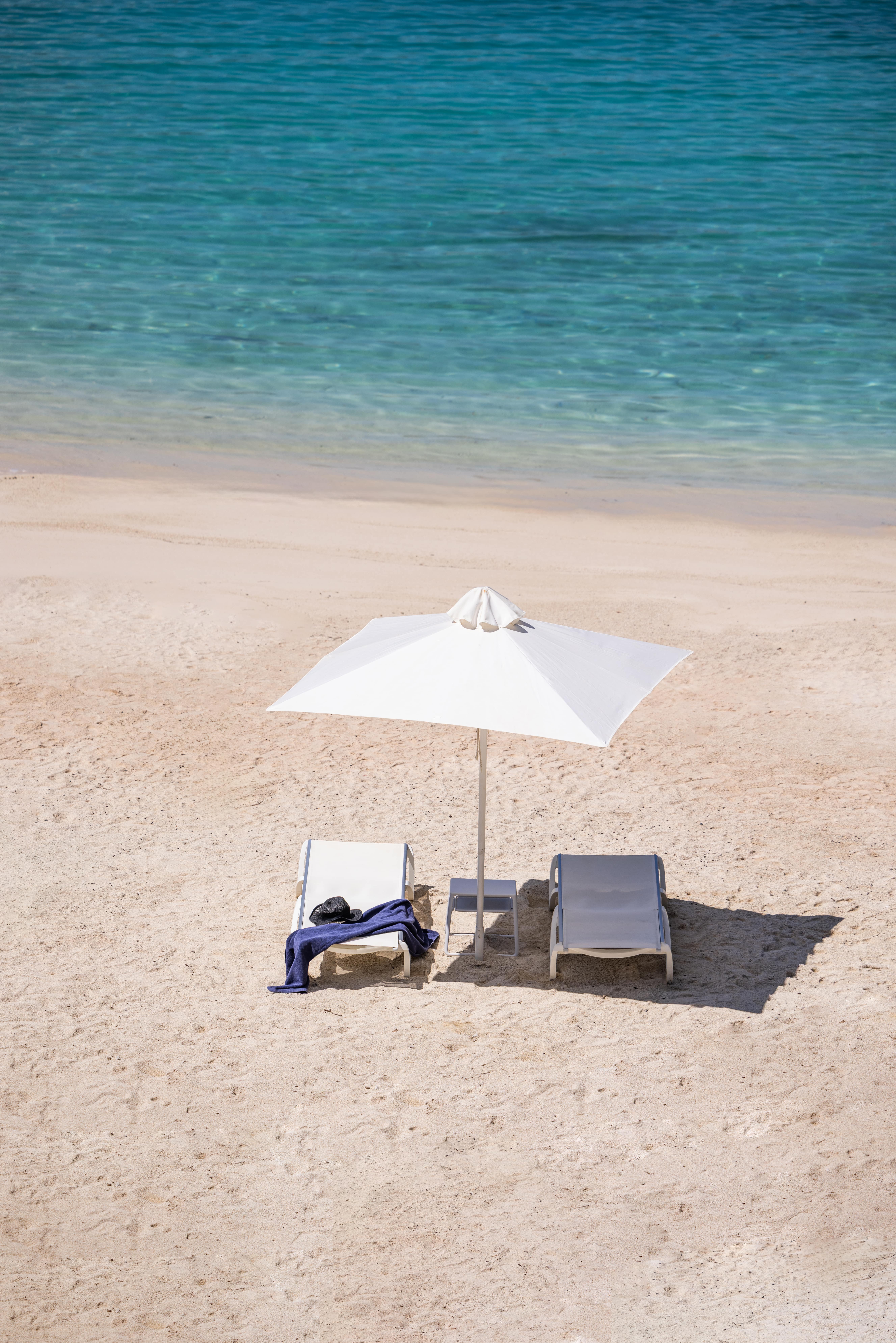 Two empty beach lounge chairs under a white umbrella on sandy shore beside clear turquoise water.