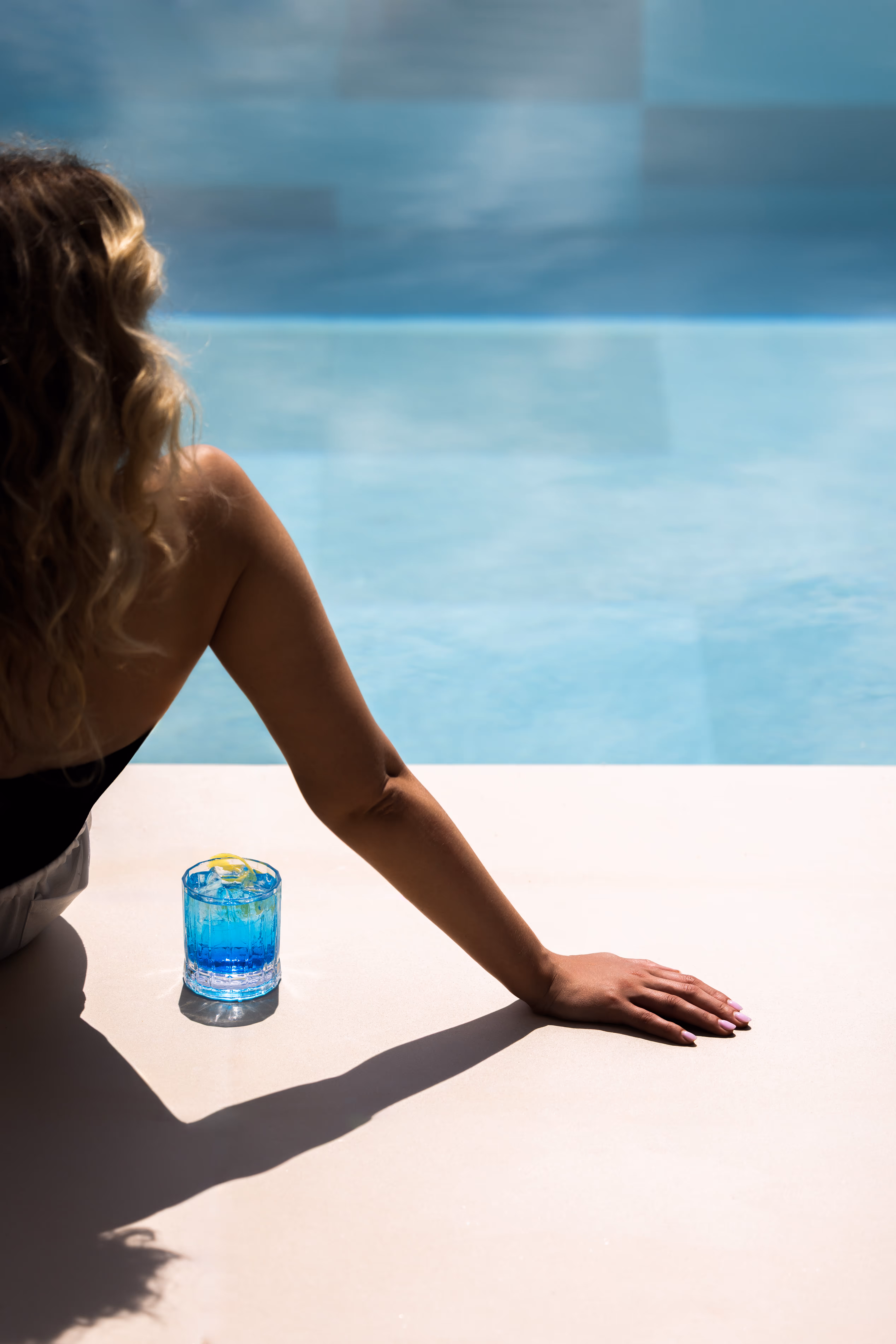 Woman with curly hair sitting by a pool edge with a blue cocktail glass beside her.