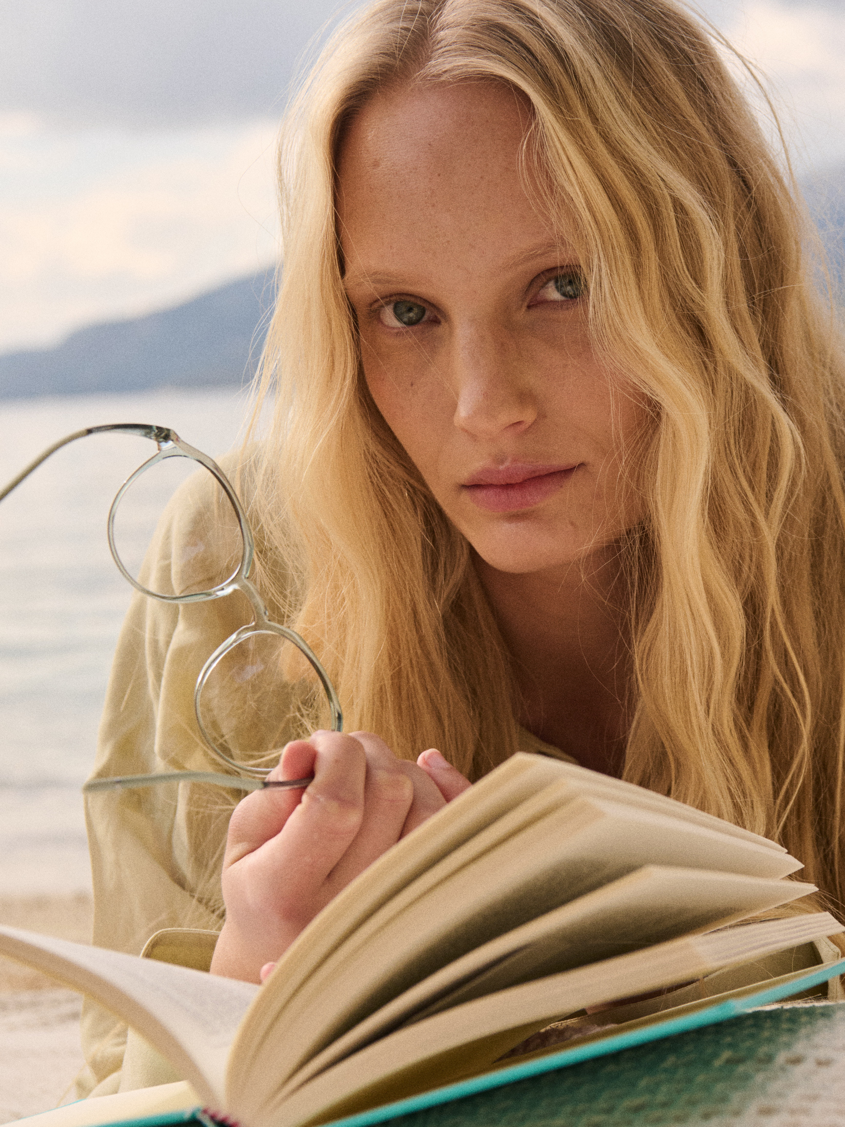 Blonde woman with wavy hair holding eyeglasses and reading a book near a calm body of water with mountains in the background.