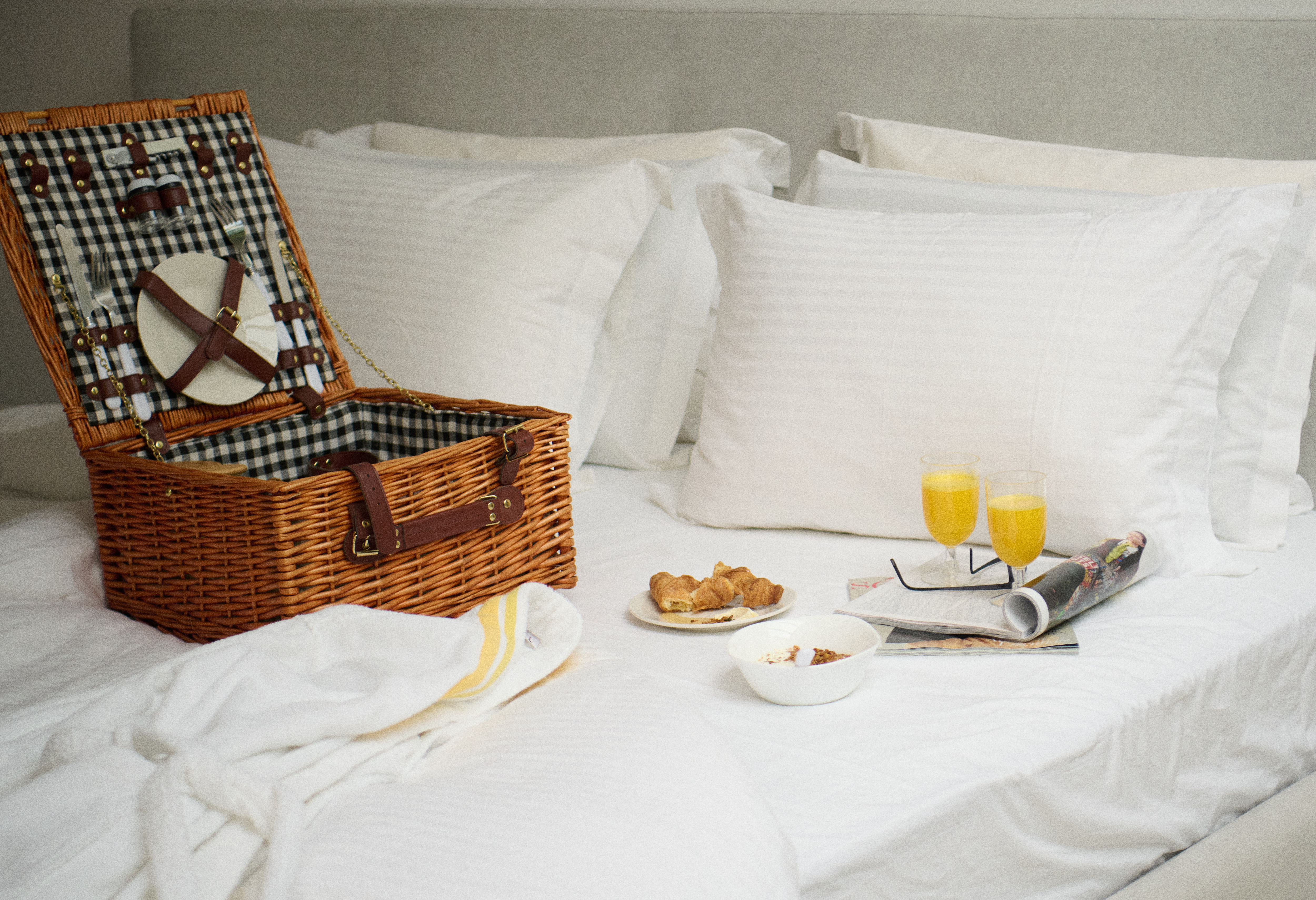 Open wicker picnic basket with plates and cutlery on a white bed next to croissants, cereal bowl, orange juice glasses, and a rolled magazine.