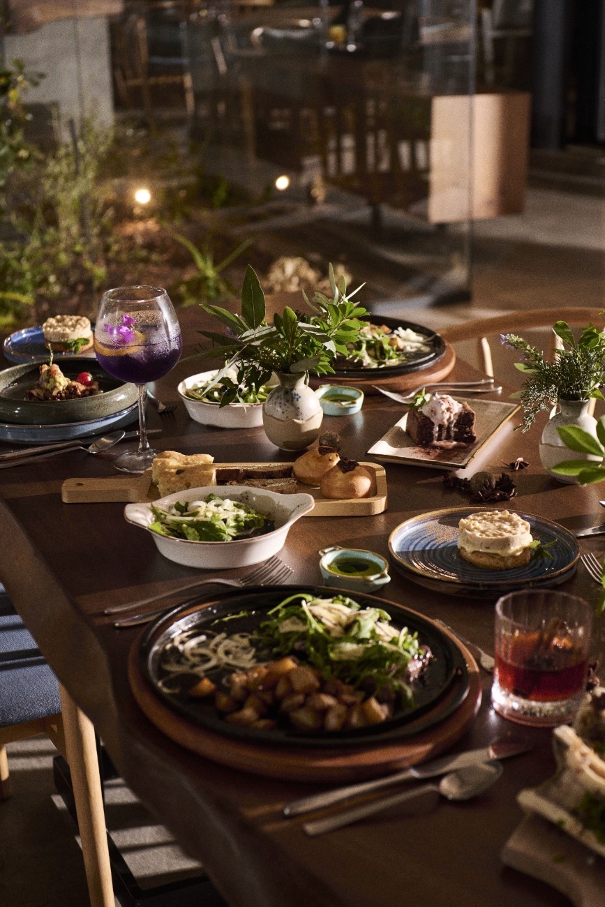 Table set with gourmet dishes including salad, bread, roasted potatoes, desserts, and vibrant cocktails in a warmly lit restaurant.