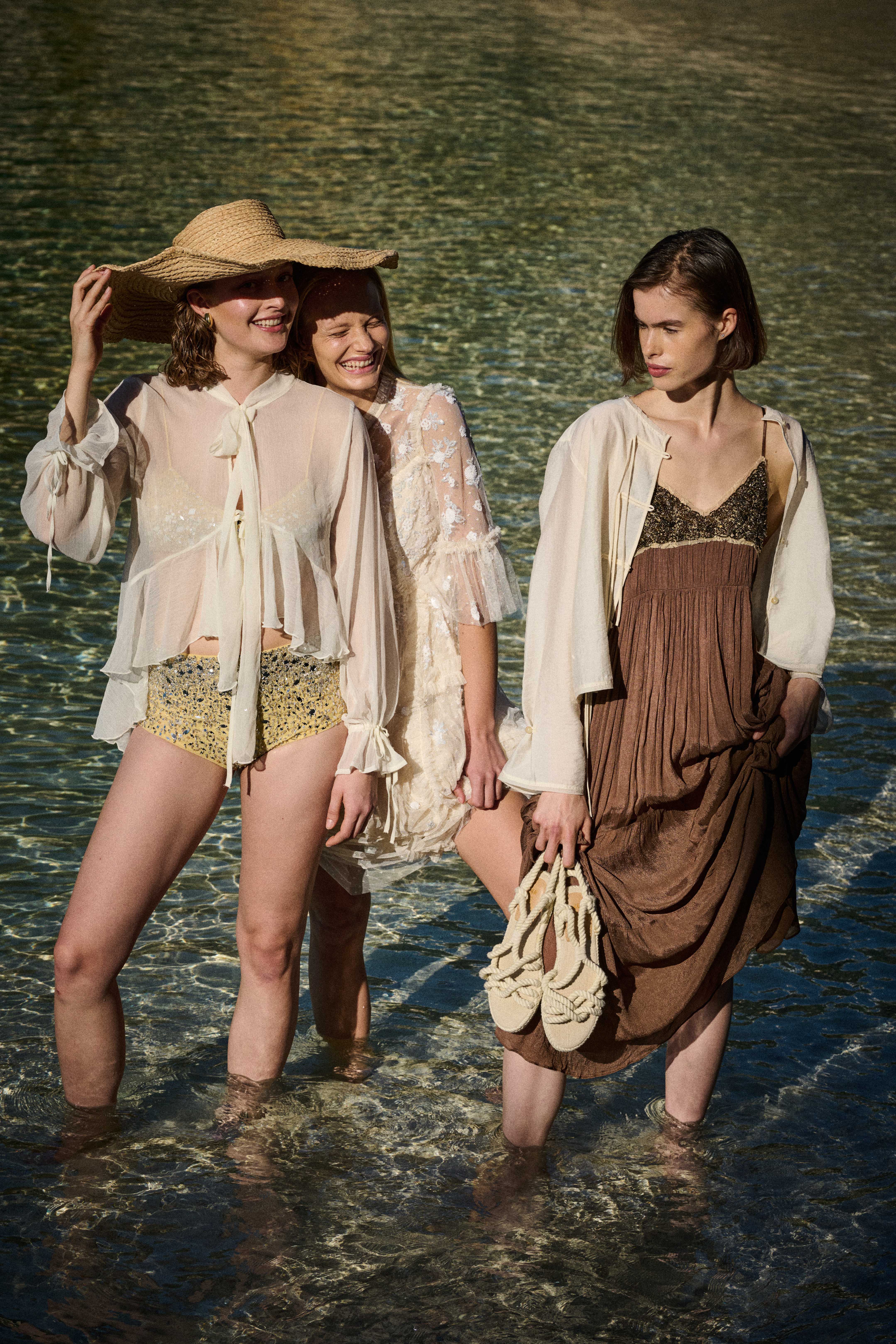 Three women standing in shallow water wearing light, flowy summer outfits; one holds a straw hat and another holds beige woven shoes.