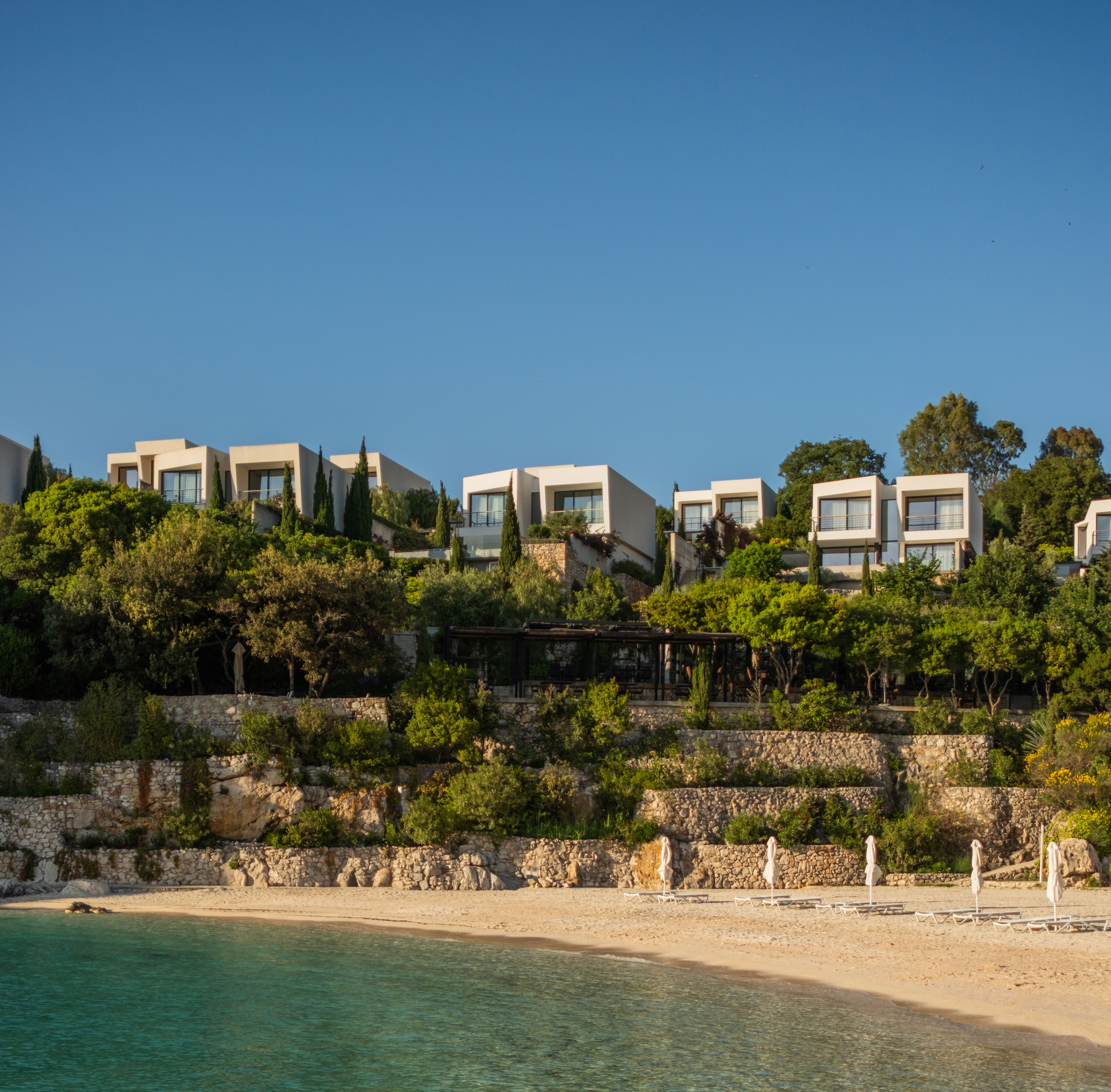 Modern white beachside villas atop terraced stone walls with green trees and umbrellas along a sandy shore by calm blue water.