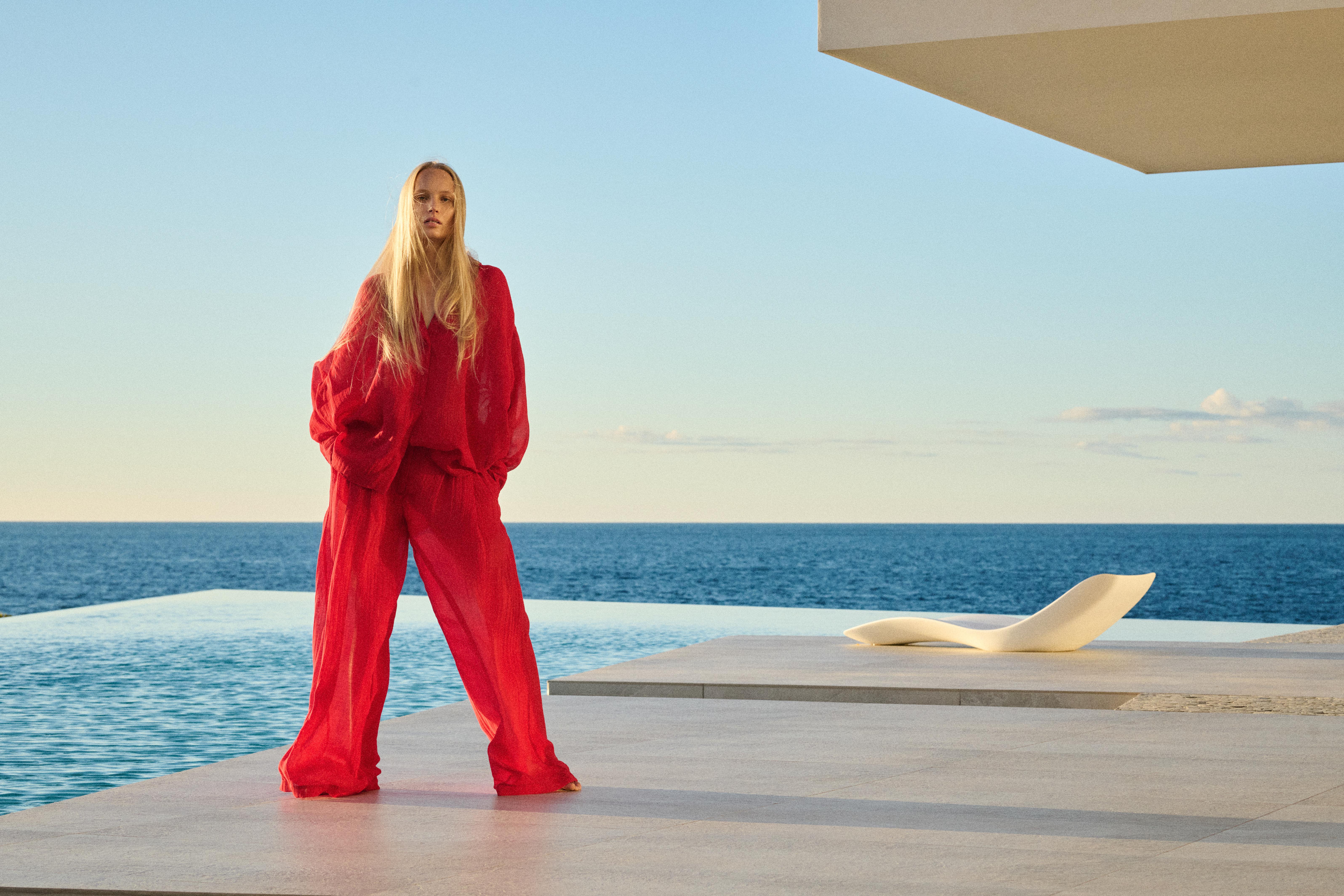 Woman with long blonde hair wearing a loose red outfit standing on a poolside terrace with ocean view and a white lounge chair.