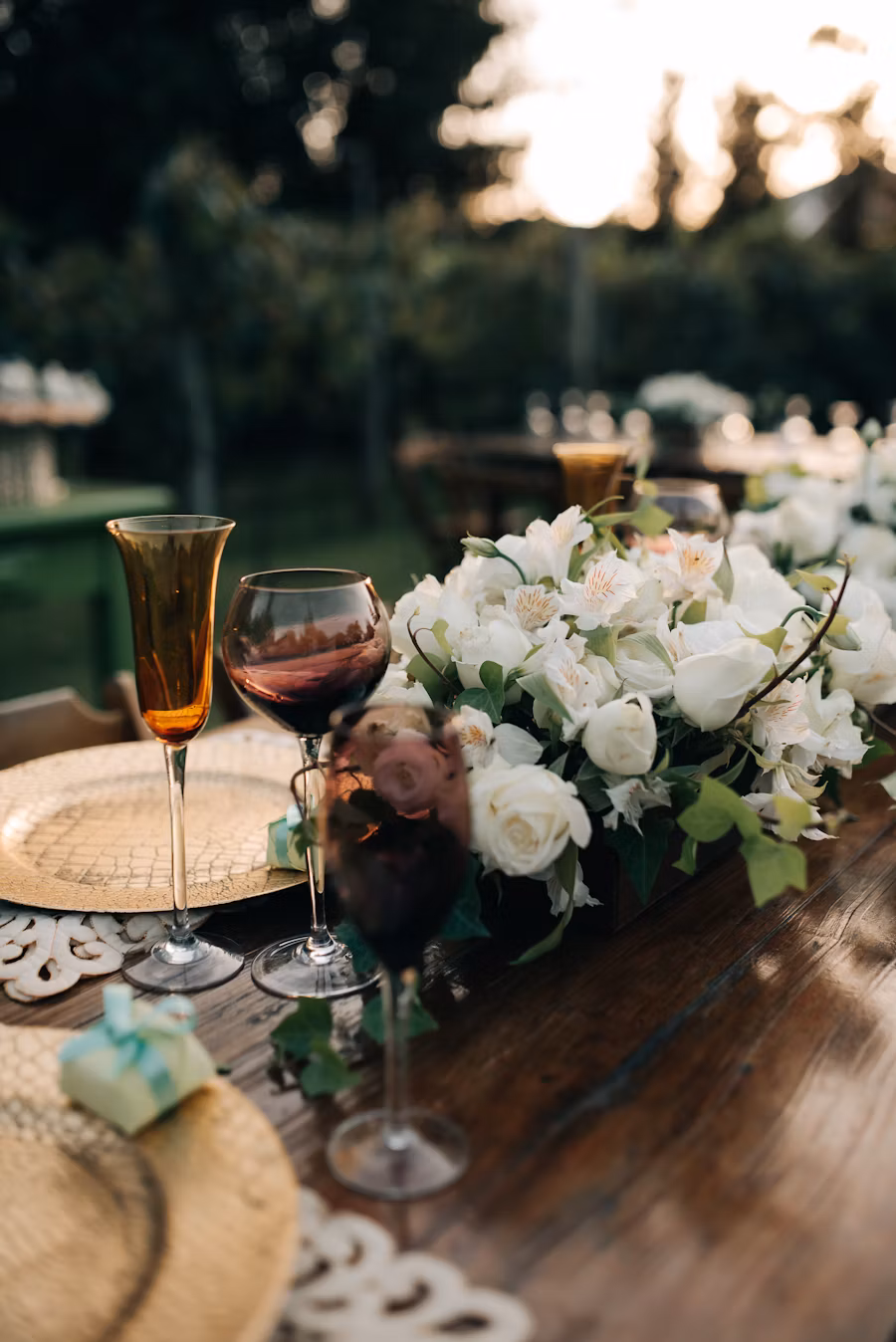 Outdoor wooden table set with gold plates, wine glasses, and a floral centerpiece of white roses and greenery.