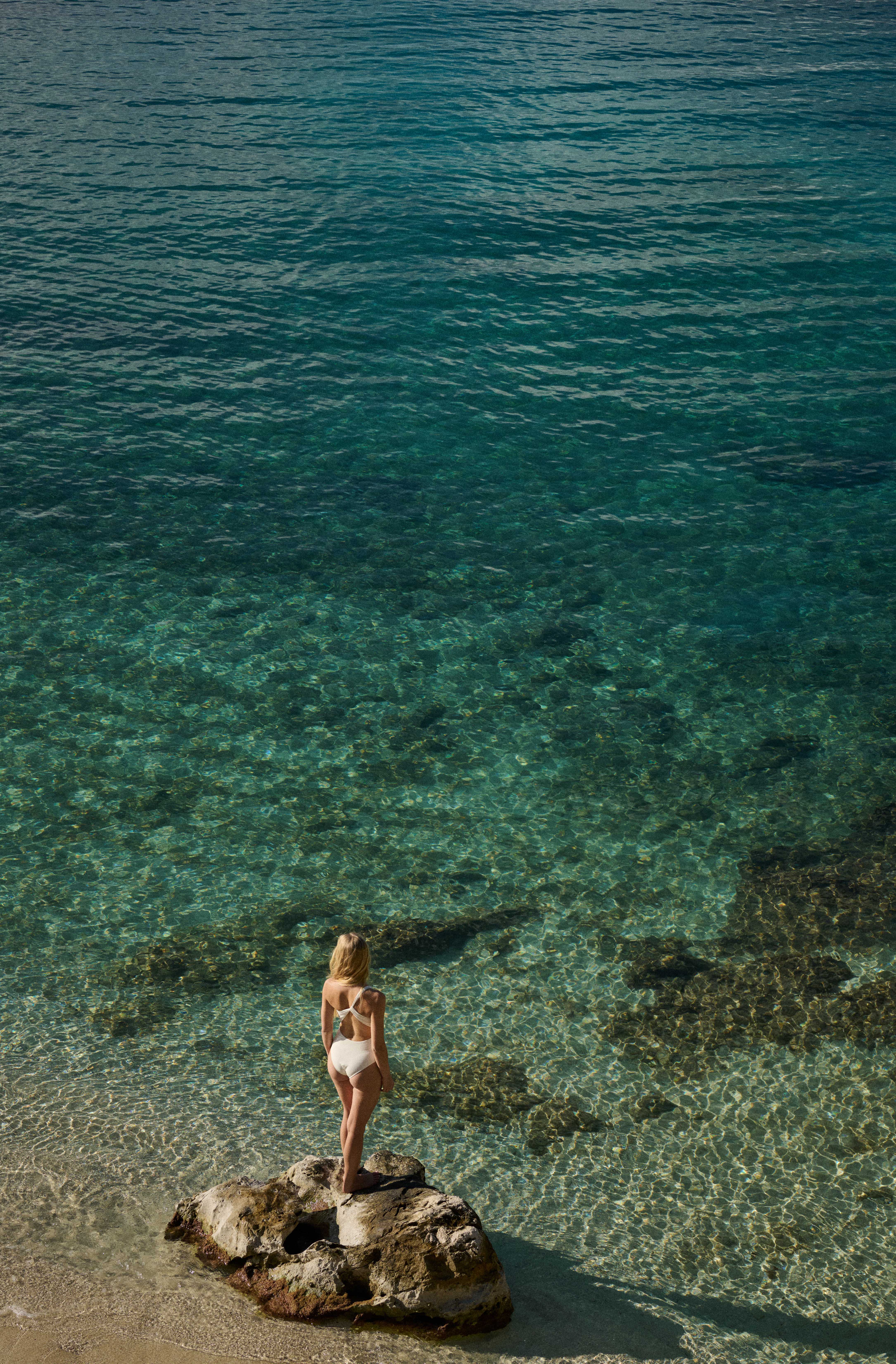 Woman in white swimsuit standing on a large rock at the edge of clear turquoise water on a sandy beach.