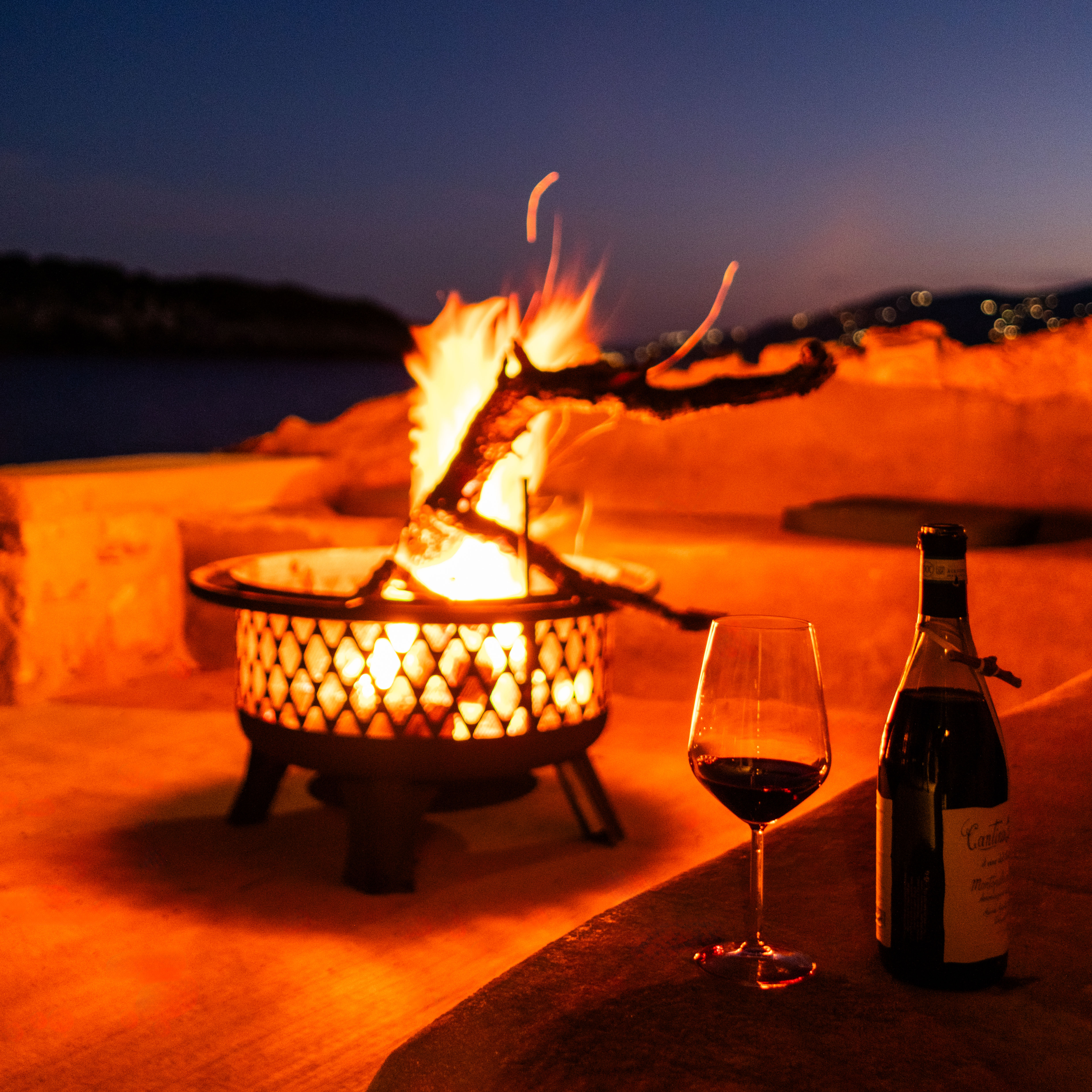 Glass of red wine and a wine bottle on a table beside a lit outdoor fire pit at dusk.