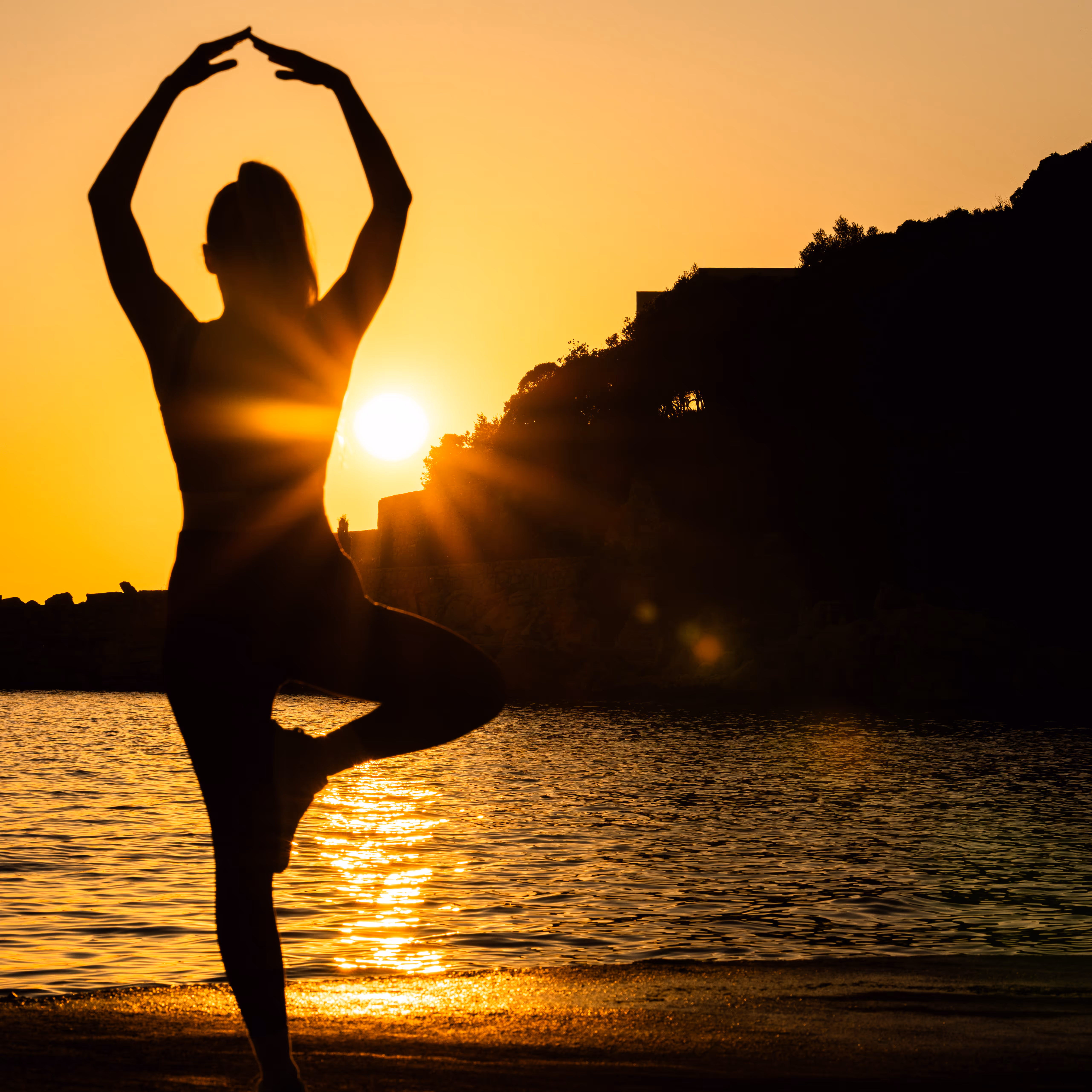 Silhouette of a person doing a yoga tree pose by the water at sunset with the sun low near the horizon.