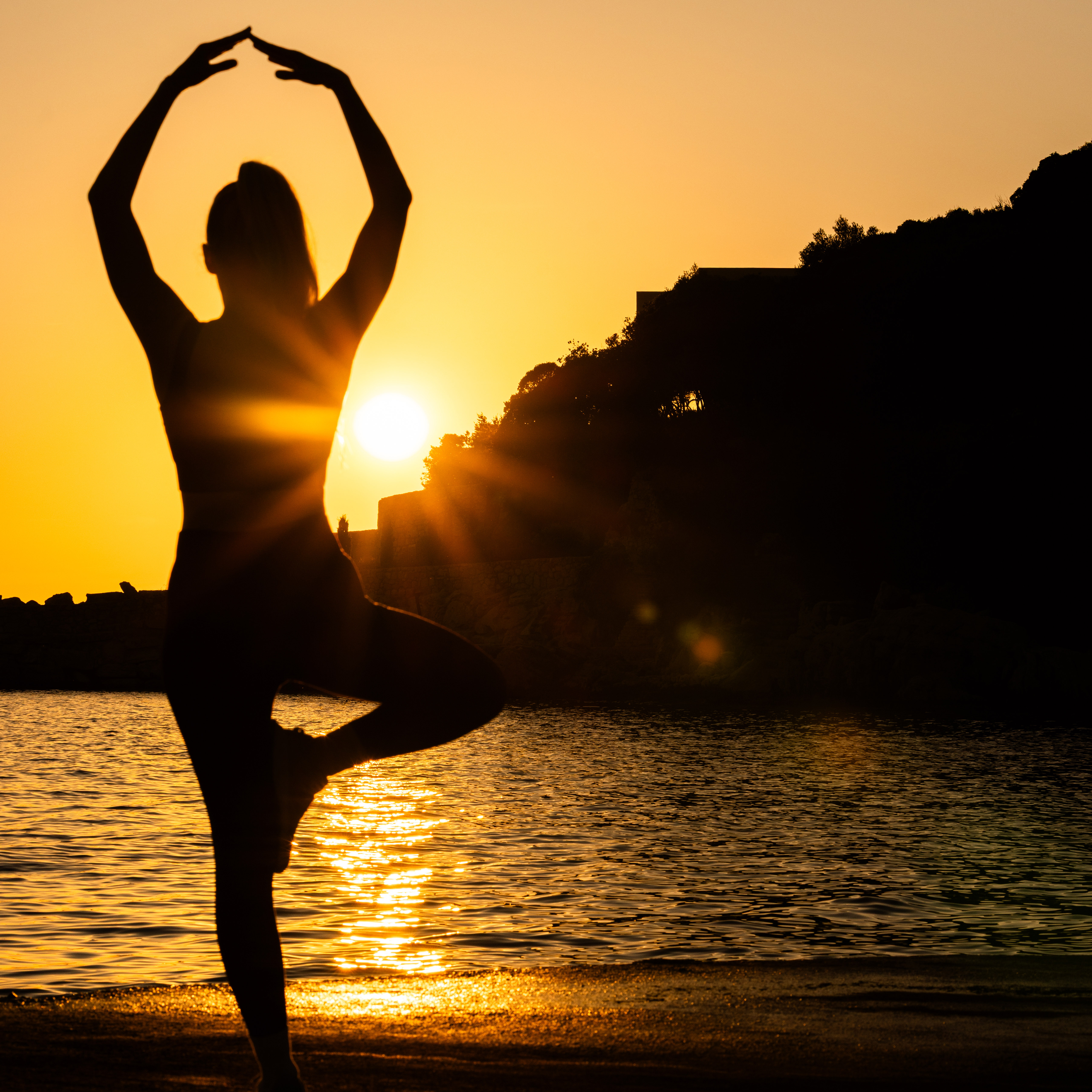 Silhouette of a person doing a yoga tree pose by the water at sunset with the sun low near the horizon.