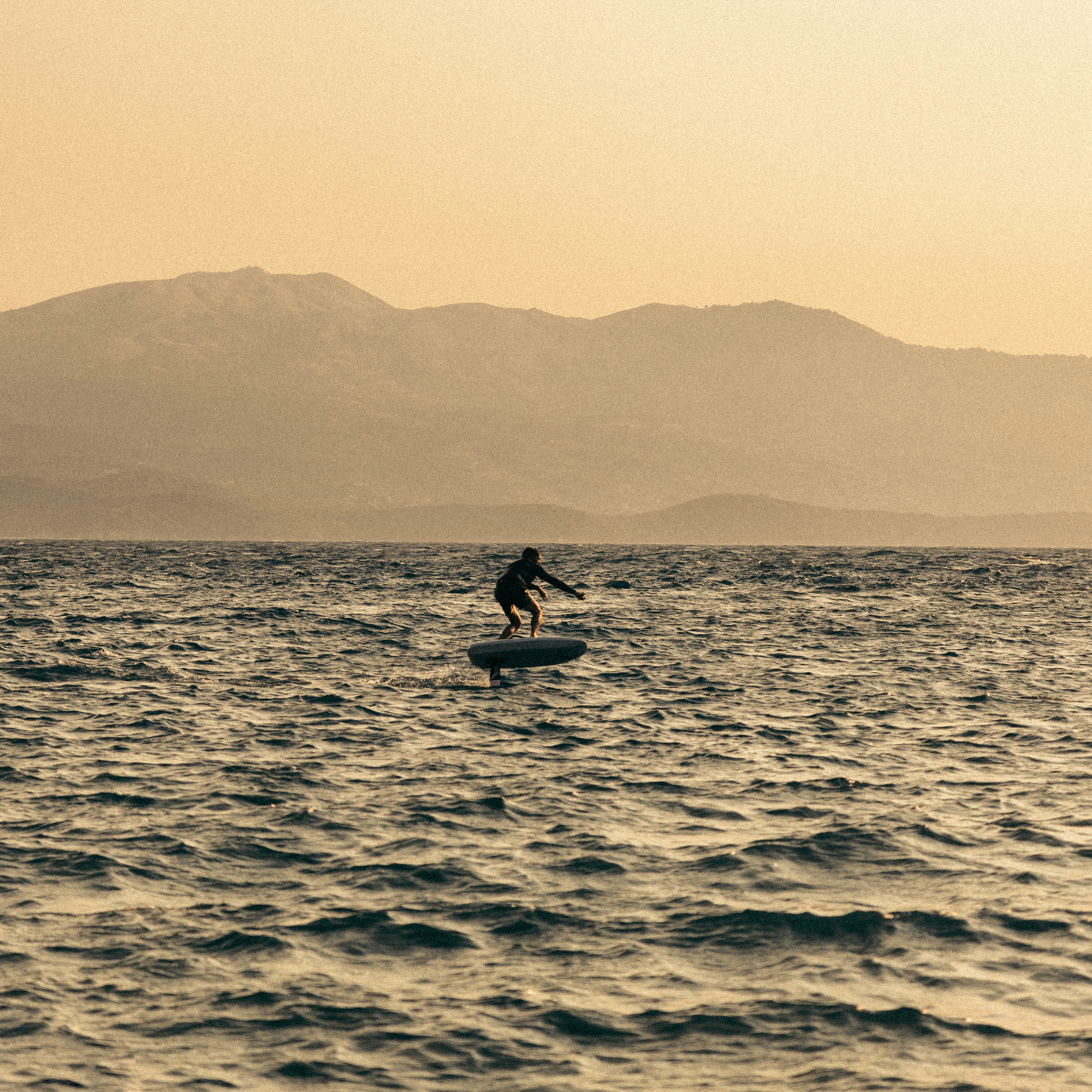 Person riding a hydrofoil surfboard on choppy sea water with mountains in the background at sunset.