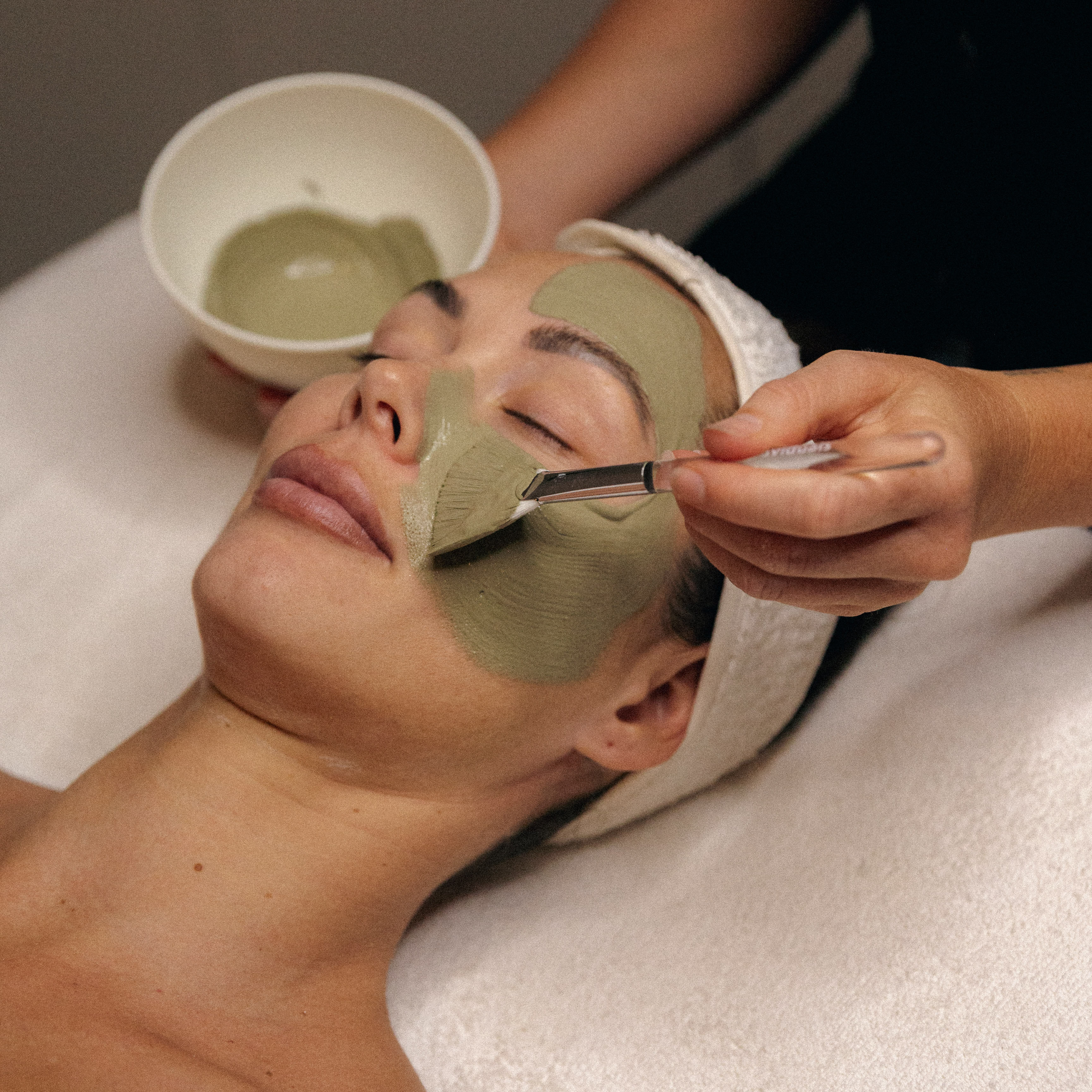 Person receiving a green facial mask applied with a brush while lying down with eyes closed and a white headband.