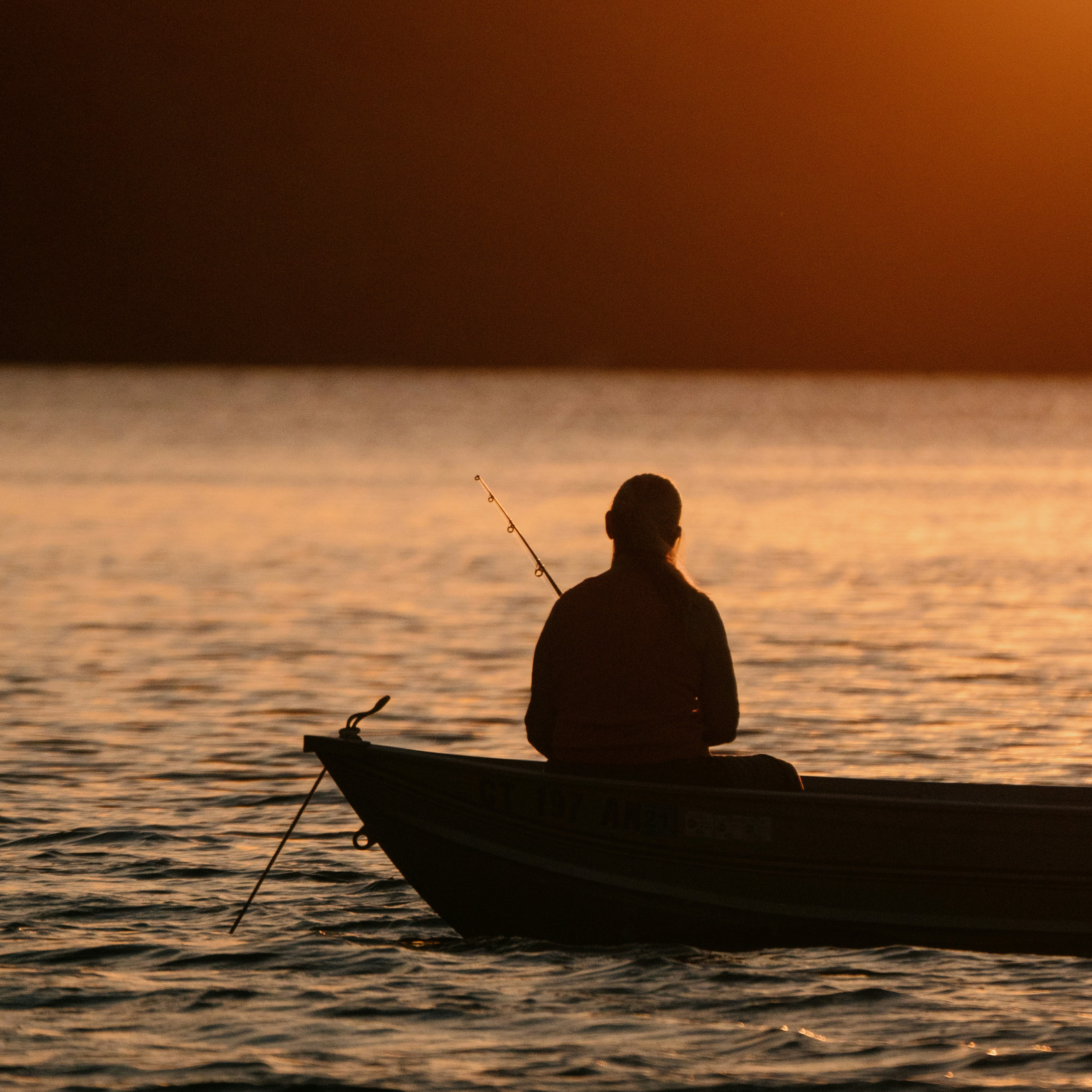 Silhouette of a person fishing from a boat on calm water during a golden sunset.