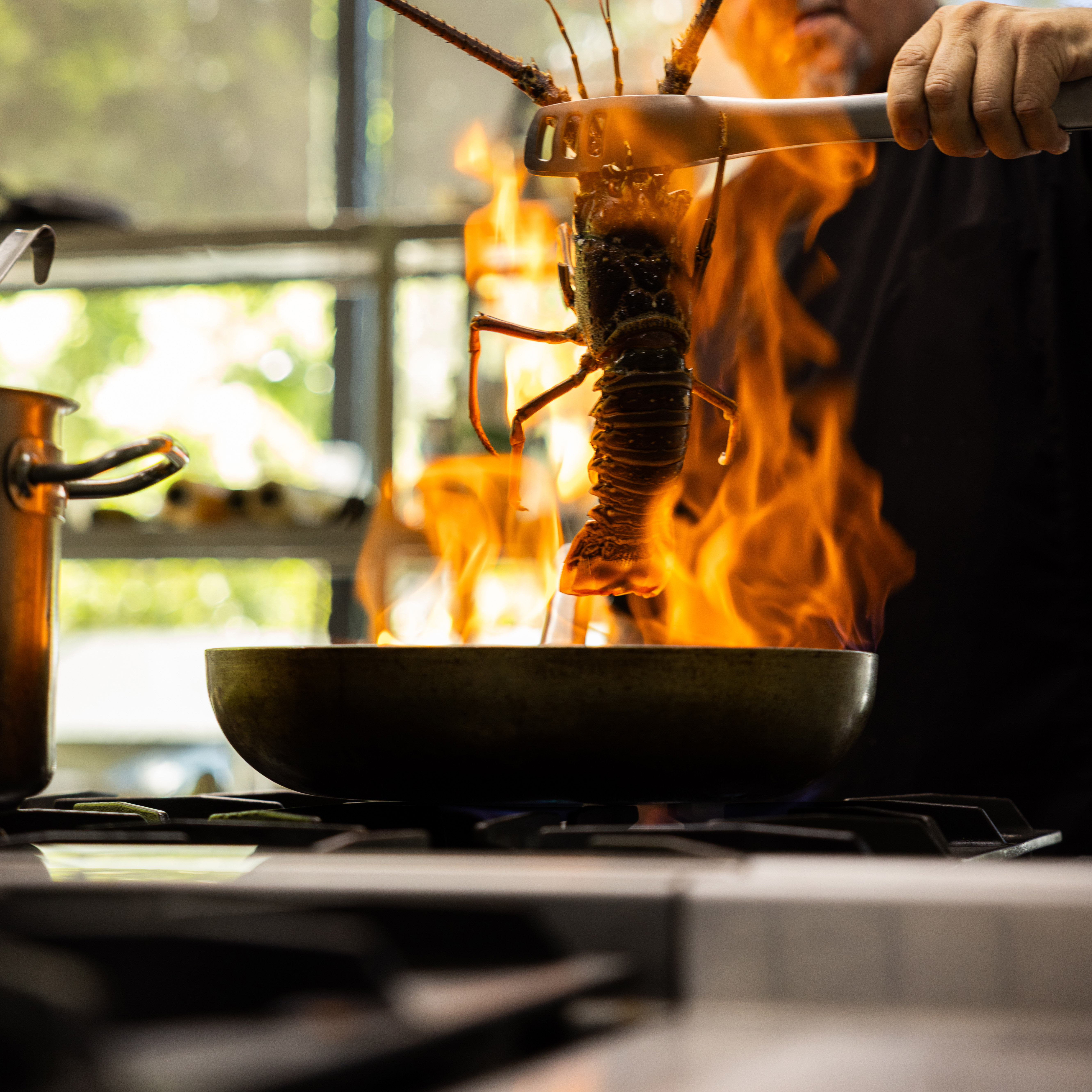 Chef holding a lobster with tongs over a flaming pan on a stove in a kitchen.